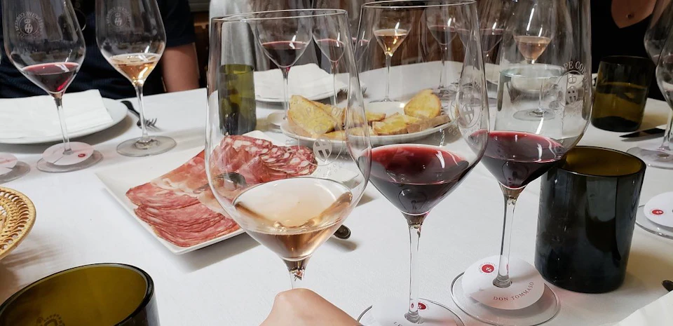Close-up of a table with wine glasses, a plate of cured meats, and a dish of sliced bread. A hand holding a wine glass in foreground.