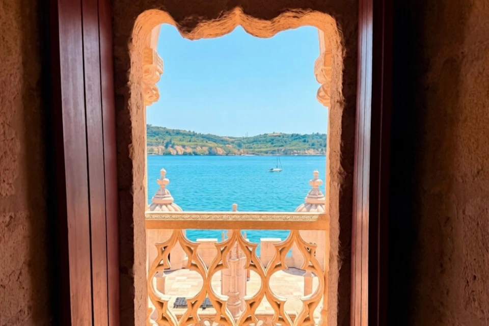 View through a decorative stone window of a balcony overlooking a calm blue sea and distant hills under a clear sky.