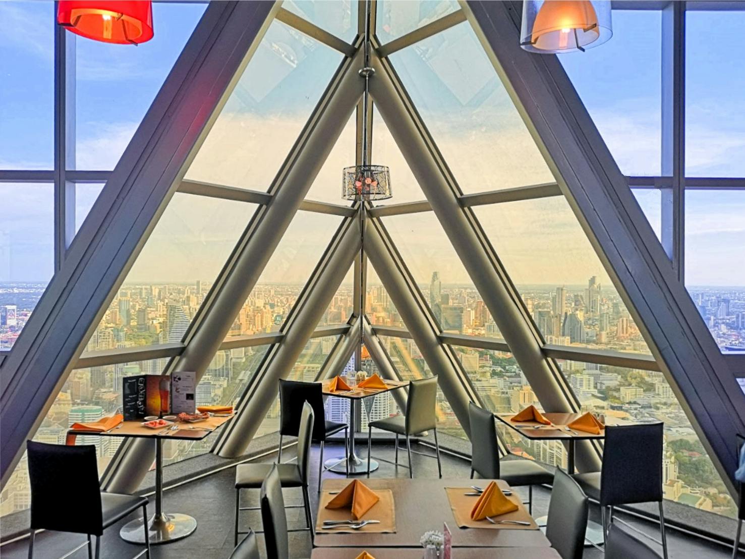 A modern restaurant with triangular glass windows offering a panoramic city view, tables set with orange napkins.