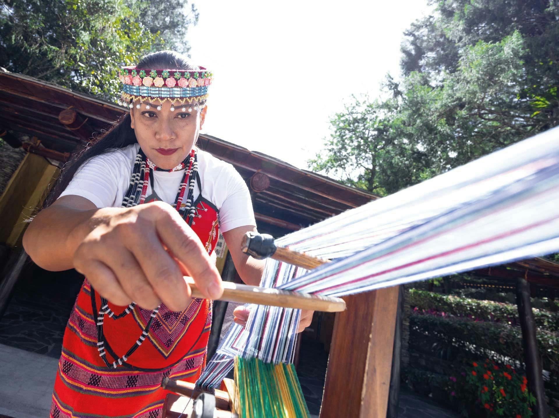 A woman wearing traditional clothing and a beaded headpiece is weaving on a loom outdoors under a sunny sky.