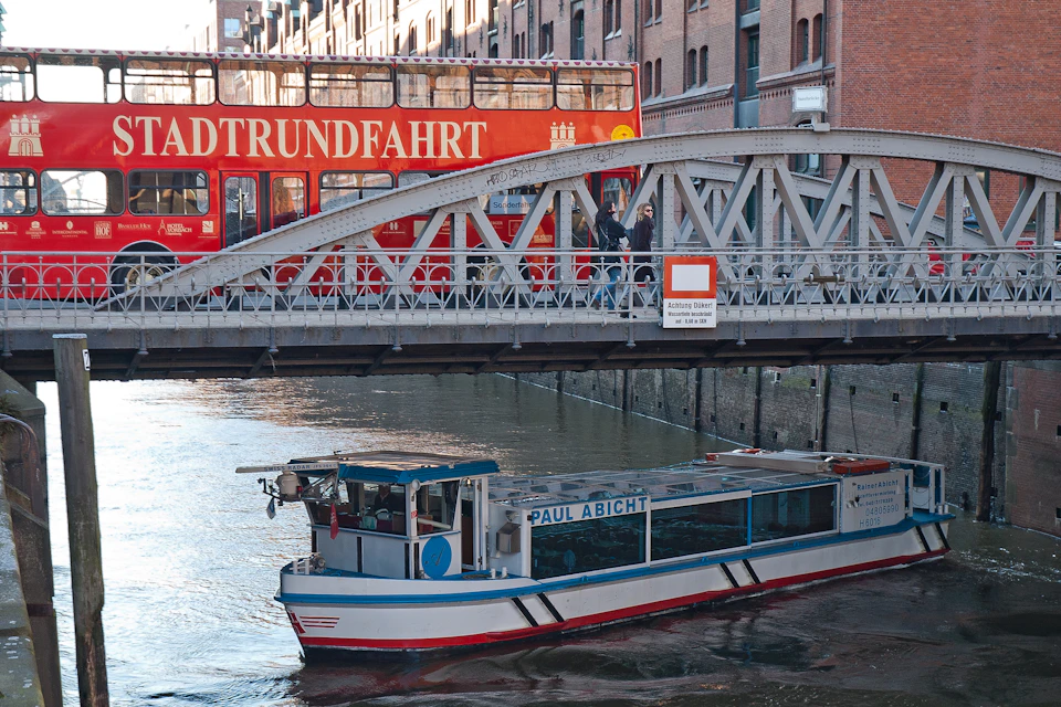 A boat on a canal passes under a bridge with pedestrians and a red double-decker bus above in a cityscape setting.