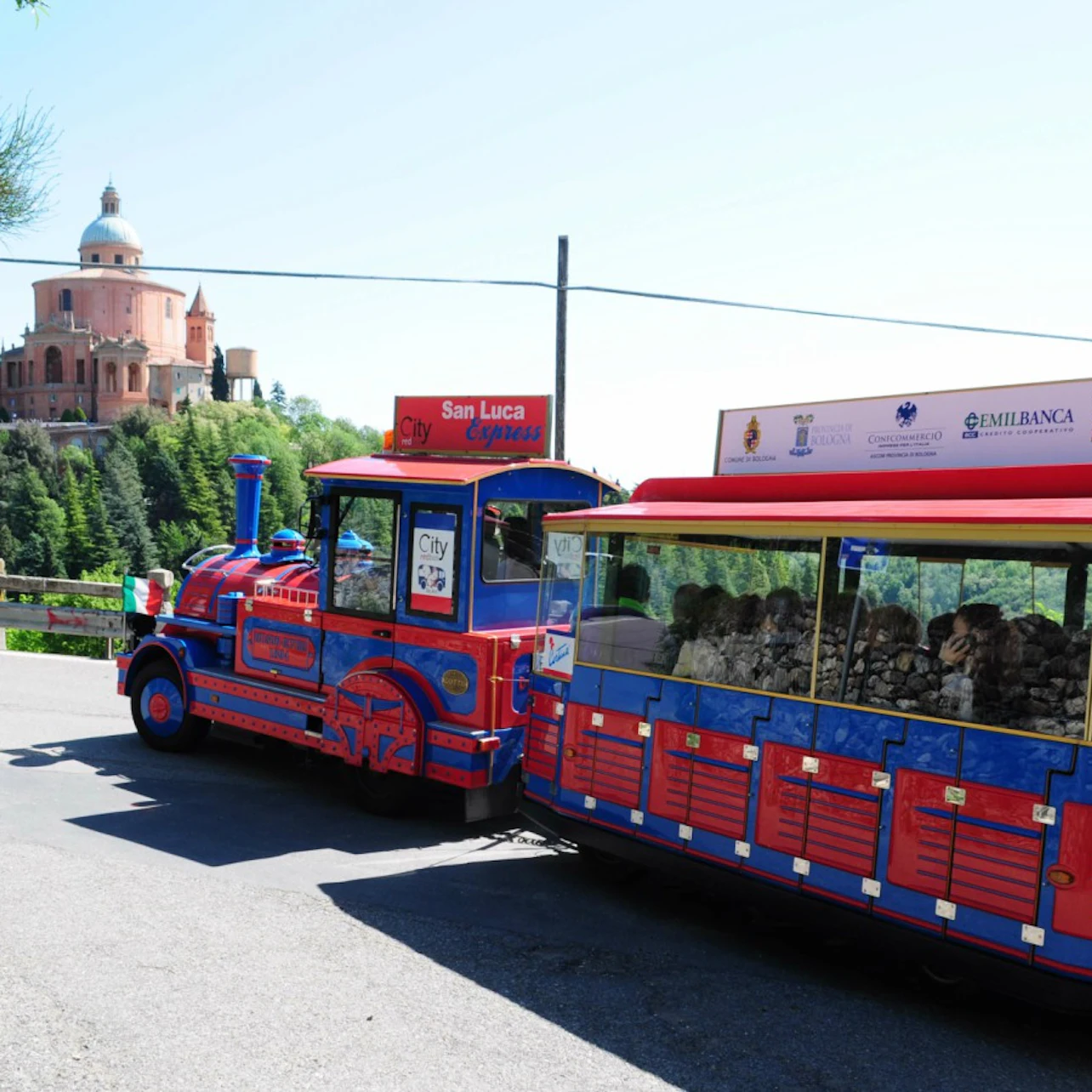 A blue and red tourist train labeled "San Luca Express" near a historic building atop a forested hill.