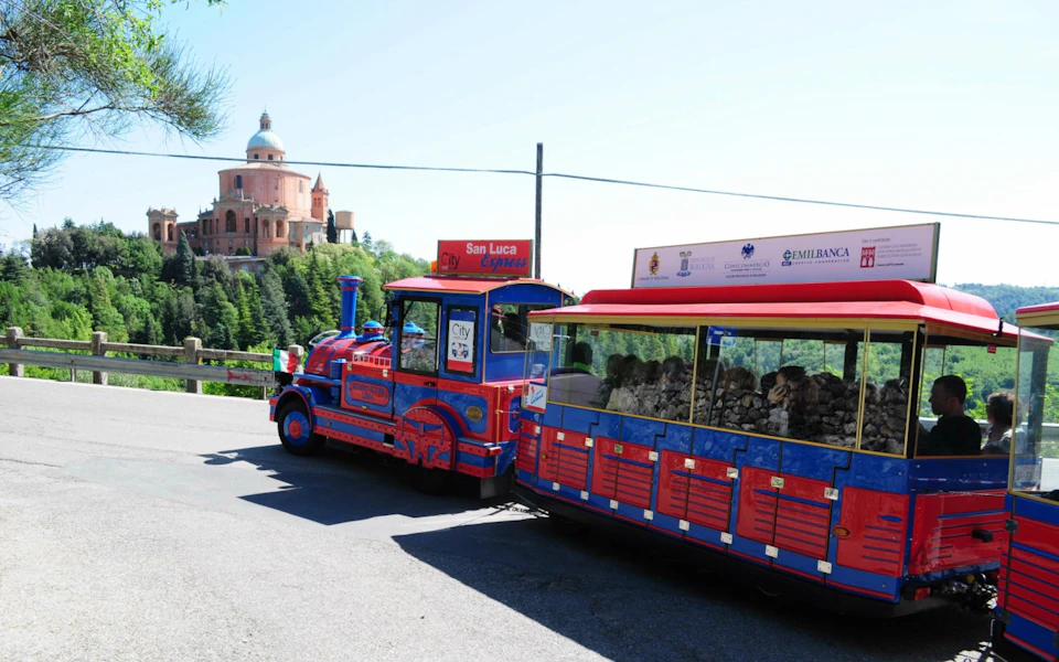 A blue and red tourist train labeled "San Luca Express" near a historic building atop a forested hill.