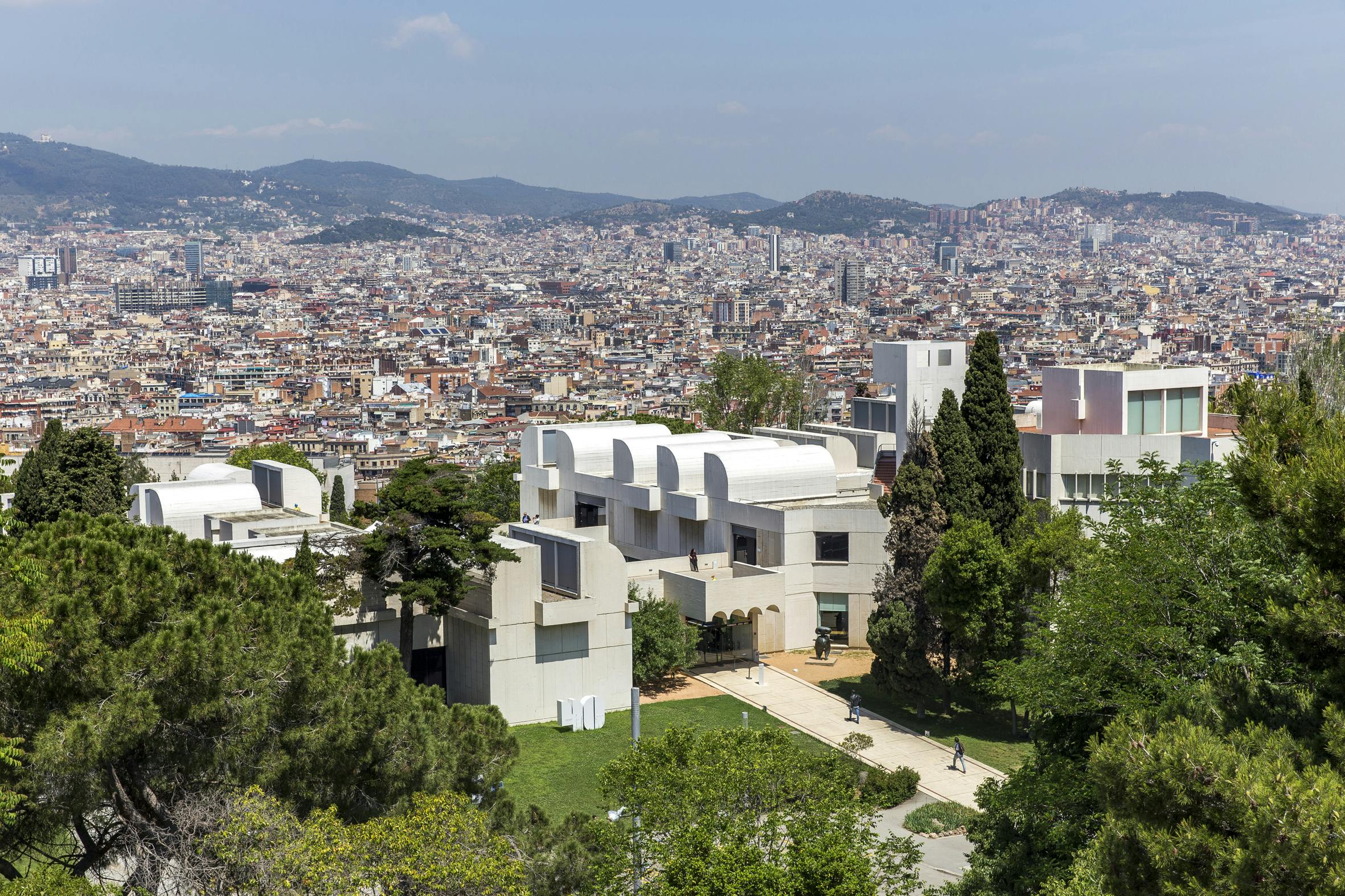 Aerial view of a modern building complex with white, curved rooftops, surrounded by greenery, and a cityscape in the background.