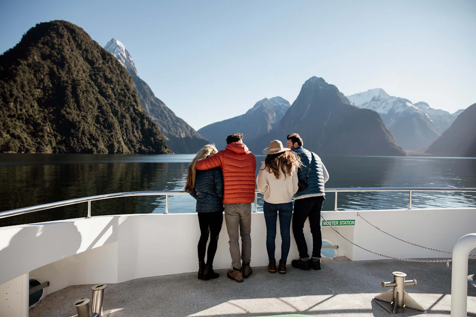 Four people standing on a boat deck, looking at a scenic fjord with tall mountains and calm water under a clear sky.