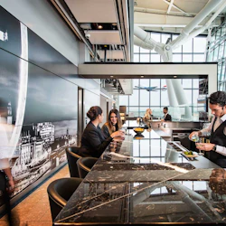Människor på en elegant flygplatsbar, en bartender som häller upp en drink, andra som sitter och pratar med ett stort fönster i bakgrunden.