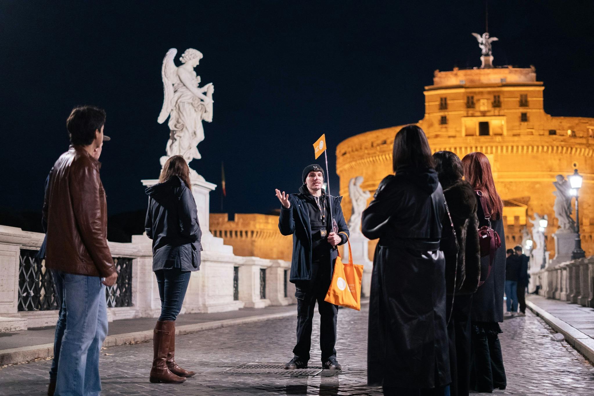 A group of people on a bridge at night near a statue, with one individual gesturing and holding a flag.