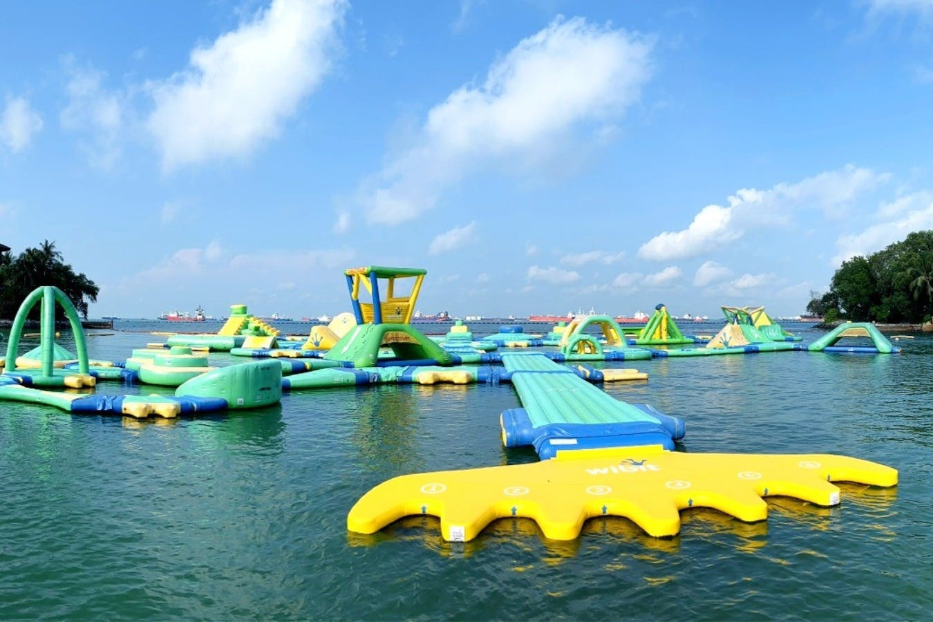 Μεγάλο πλωτό φουσκωτό υδάτινο πάρκο με τσουλήθρες και εμπόδια μια ηλιόλουστη μέρα, περιτριγυρισμένο από ήρεμα νερά και καταγάλανο ουρανό.