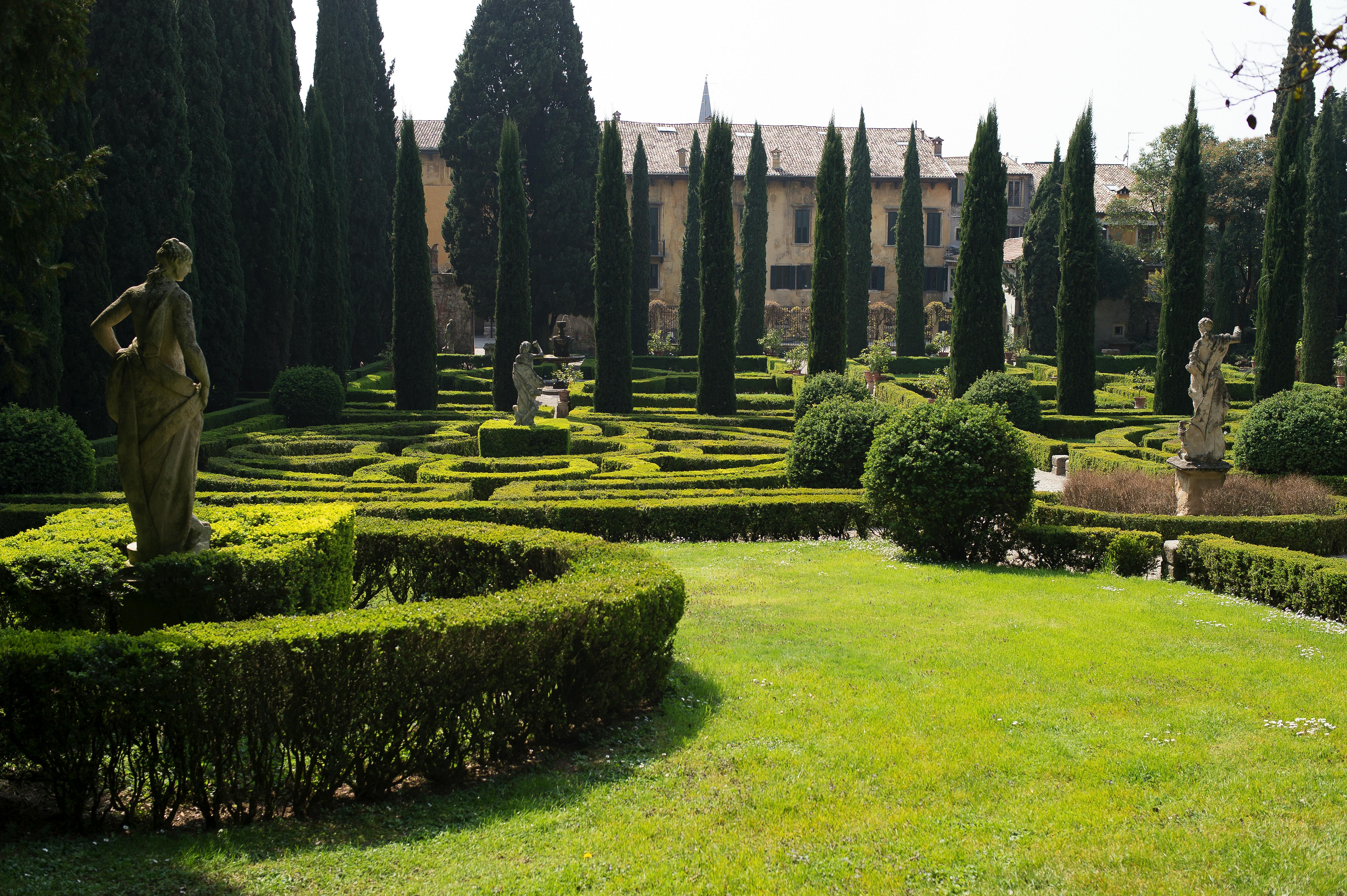 精心修剪的花园，高大的柏树，树篱形成的图案，远处的雕像，以及后面的历史建筑。