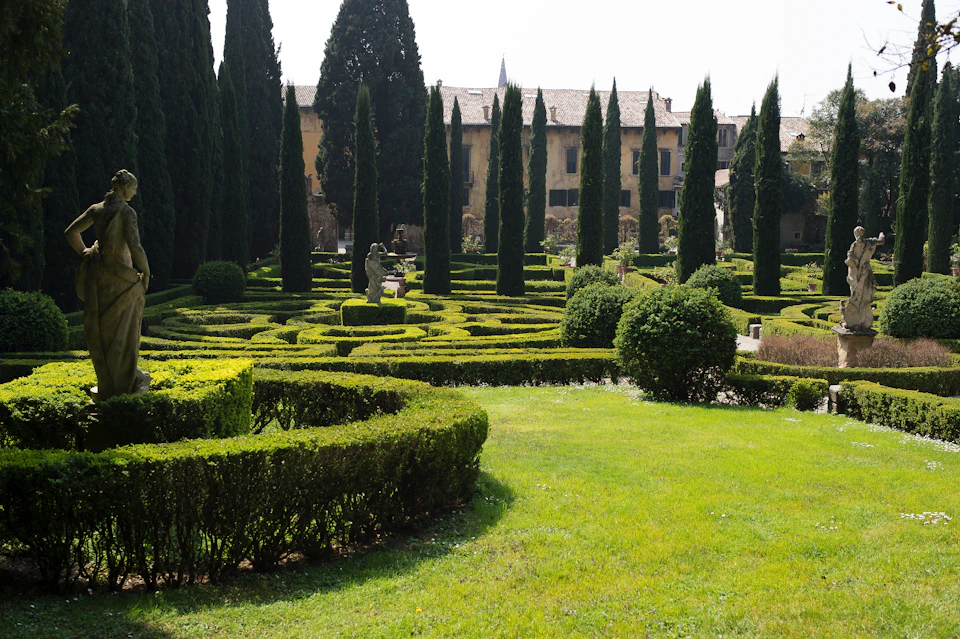 Well-manicured garden with tall cypress trees, hedges forming patterns, a statue in the distance, and historic buildings behind.