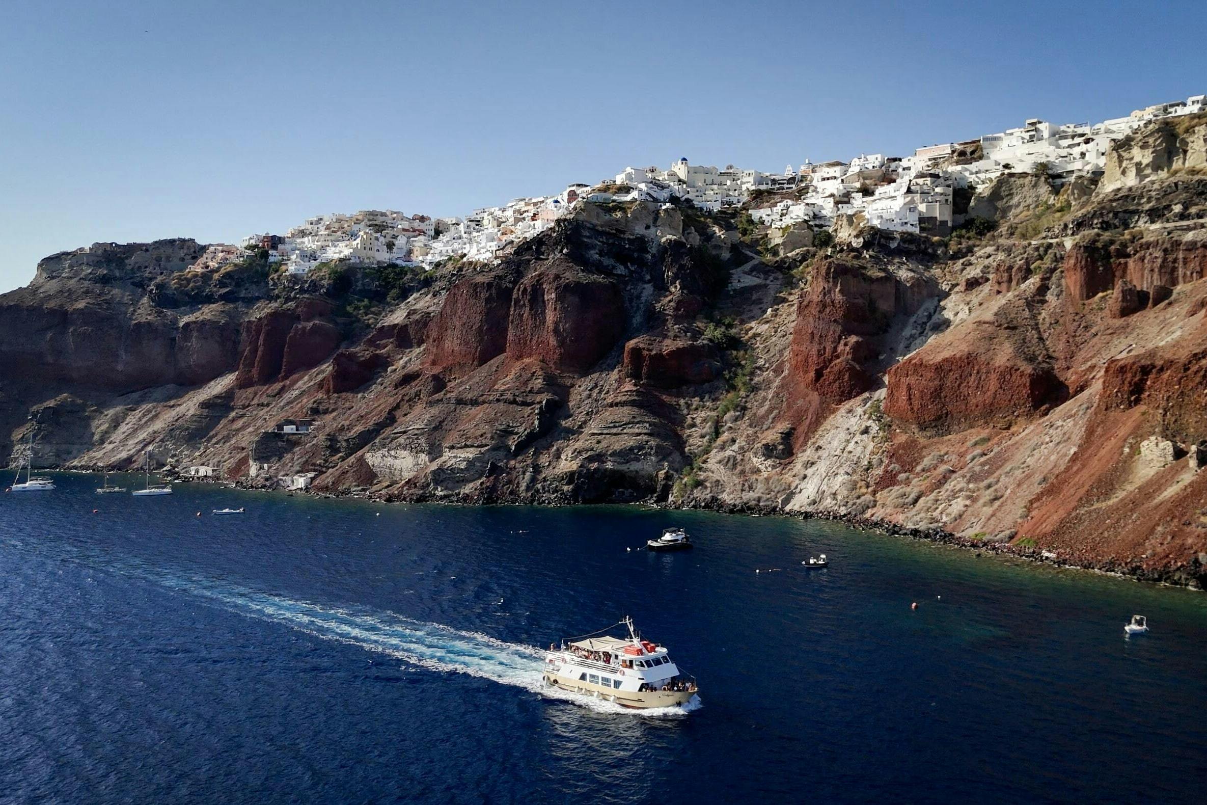 Ένας παραθαλάσσιος βράχος με λευκά σπίτια στην κορυφή, μια βάρκα που πλοηγεί στη γαλάζια θάλασσα από κάτω και μικρότερα σκάφη διάσπαρτα κοντά.