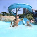 Due ragazzi giocano e sguazzano in una piscina sotto un grande scivolo blu in una giornata di sole.