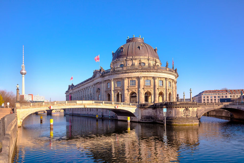A historic building with a dome and flags, next to a bridge over a river, under a clear blue sky.