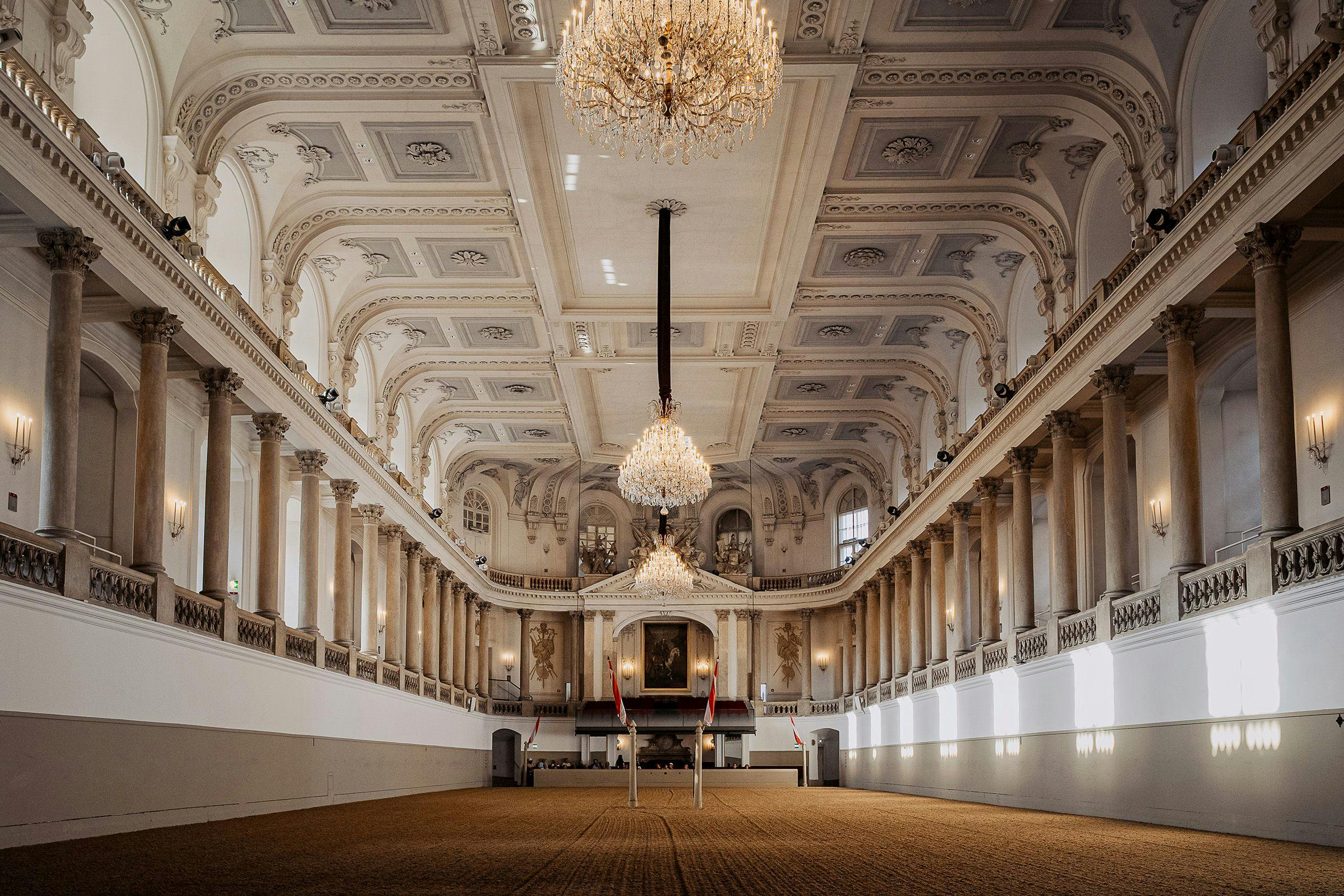 Large ornate hall with intricate ceilings, chandeliers, and a stage with flags. Pillars line both sides, and the hall is well-lit.