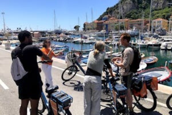 Four people with bicycles stand by a marina with docked boats, having a conversation on a sunny day.