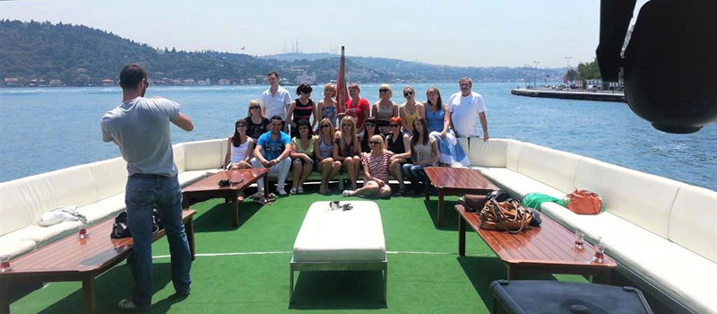 A group of people posing on a boat with white seating and green carpet, against a backdrop of water and distant hills.