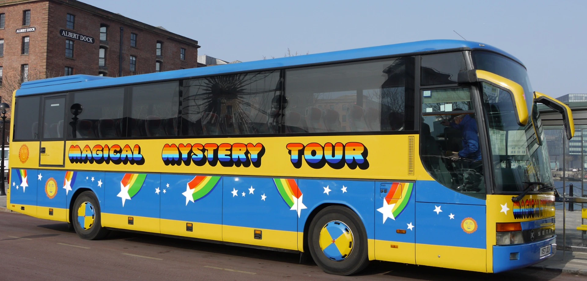 A colorful bus with "Mystery Tour" written on its side, adorned with rainbows and stars, parked in an urban area.