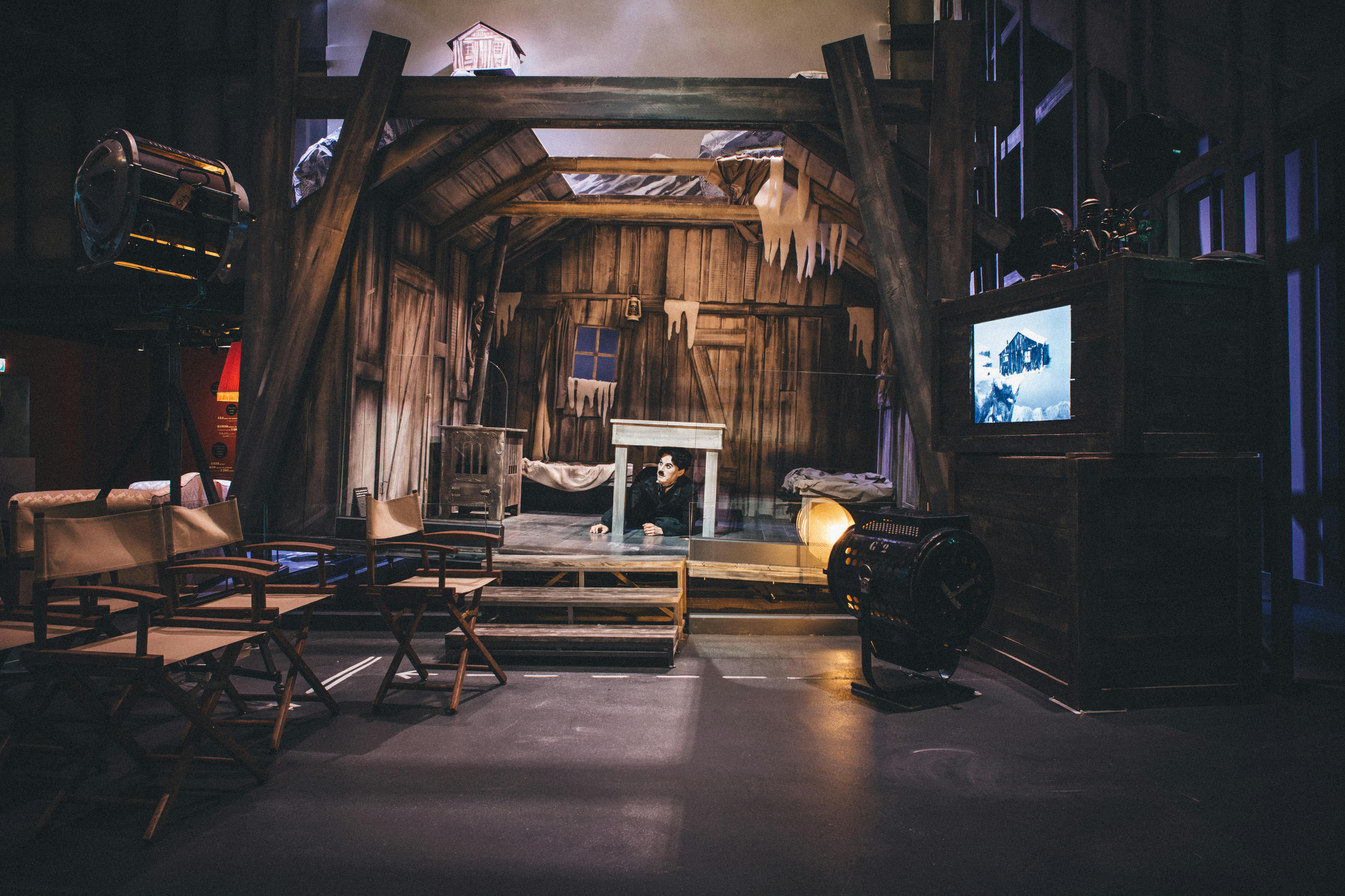 Rustikální scéna připomínající starý dřevěný dům s rampouchy. Pod stolem vykukuje herec. Filmové vybavení a židle jsou vidět.