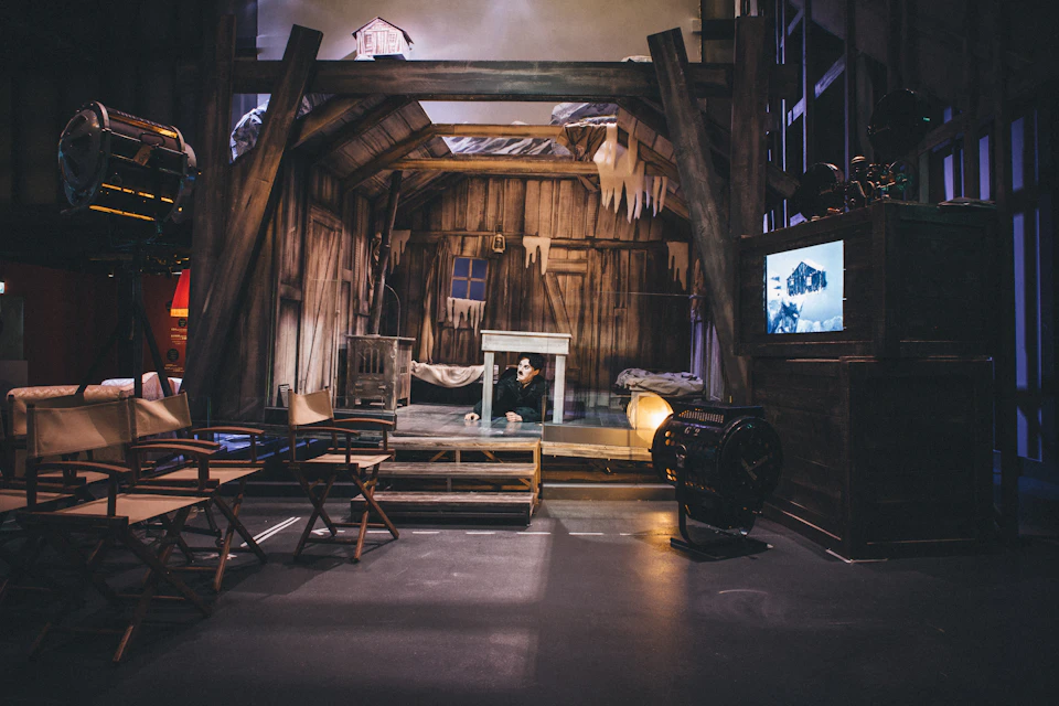 A rustic stage set resembling an old, wooden house with icicles. An actor peeks from under a table. Film equipment and chairs are seen.