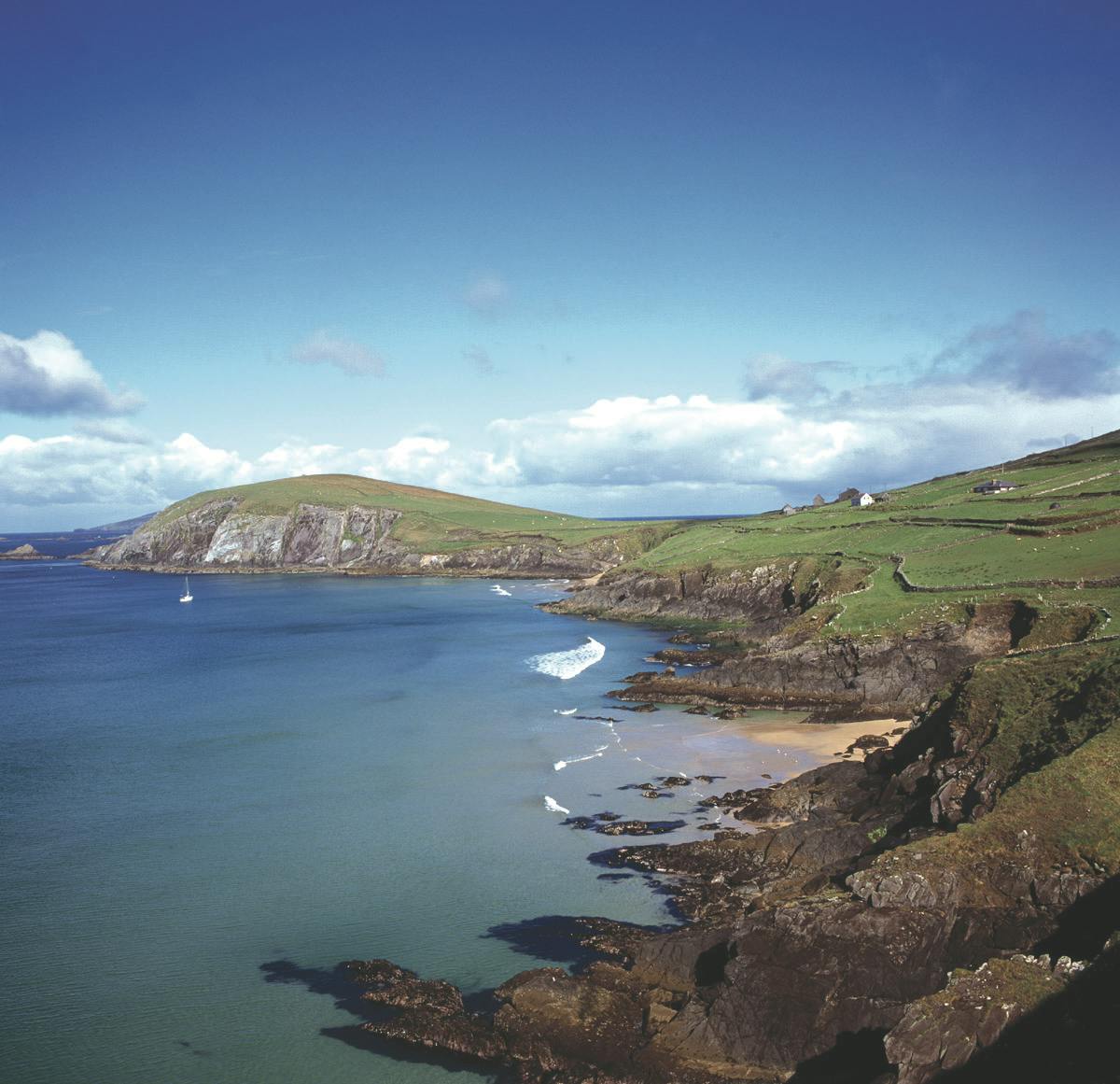 A rocky coastline with green hills, waves crashing on the shore, a distant sailboat, and a clear blue sky.