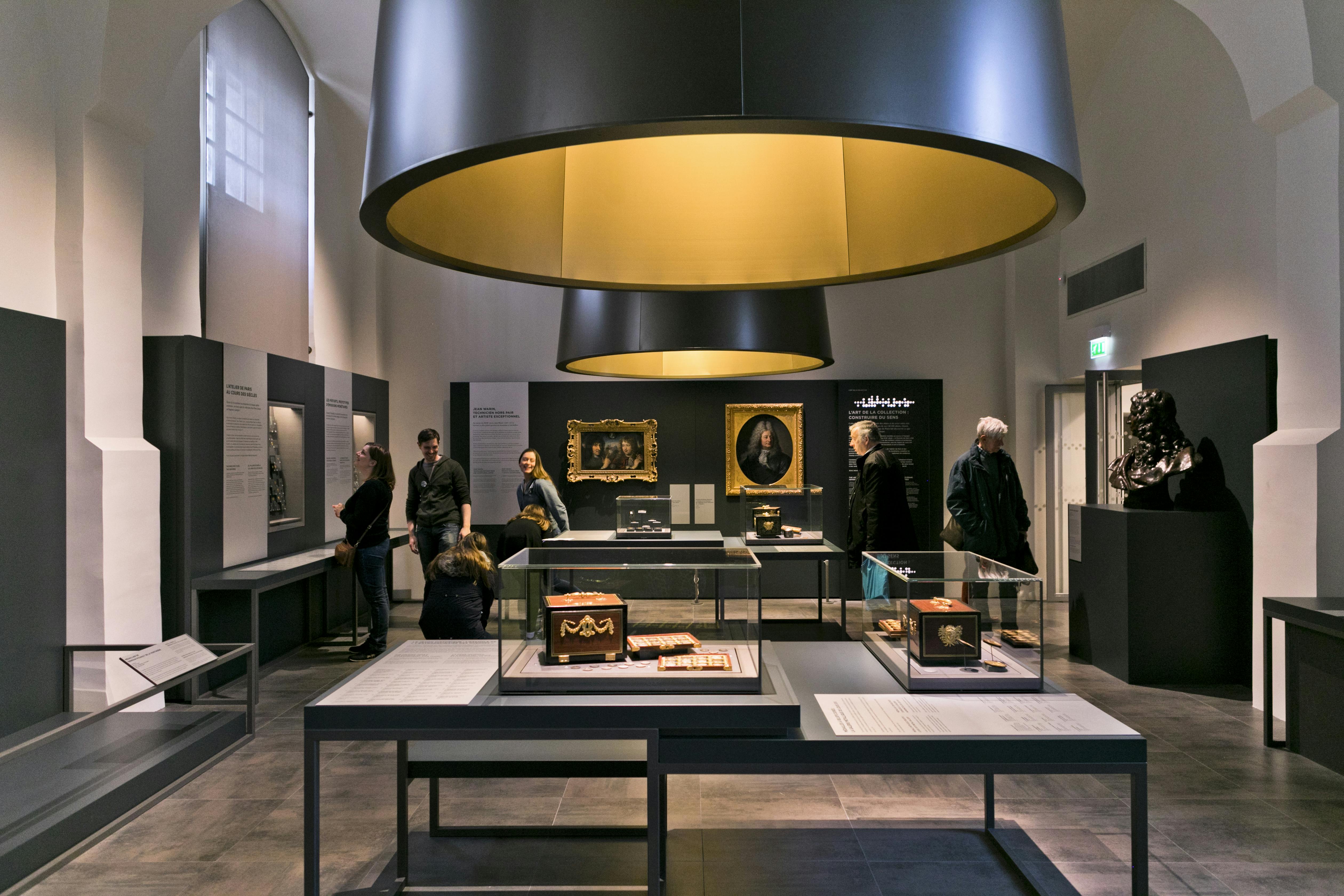 Άνθρωποι που βλέπουν ιστορικά αντικείμενα και πίνακες ζωγραφικής που εκτίθενται σε μια καλά φωτισμένη μουσειακή γκαλερί με μεγάλα κυκλικά φώτα.