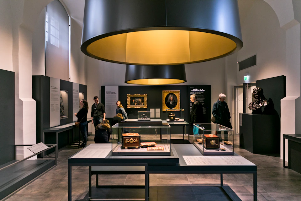 People viewing historical artifacts and paintings displayed in a well-lit museum gallery with large circular overhead lights.