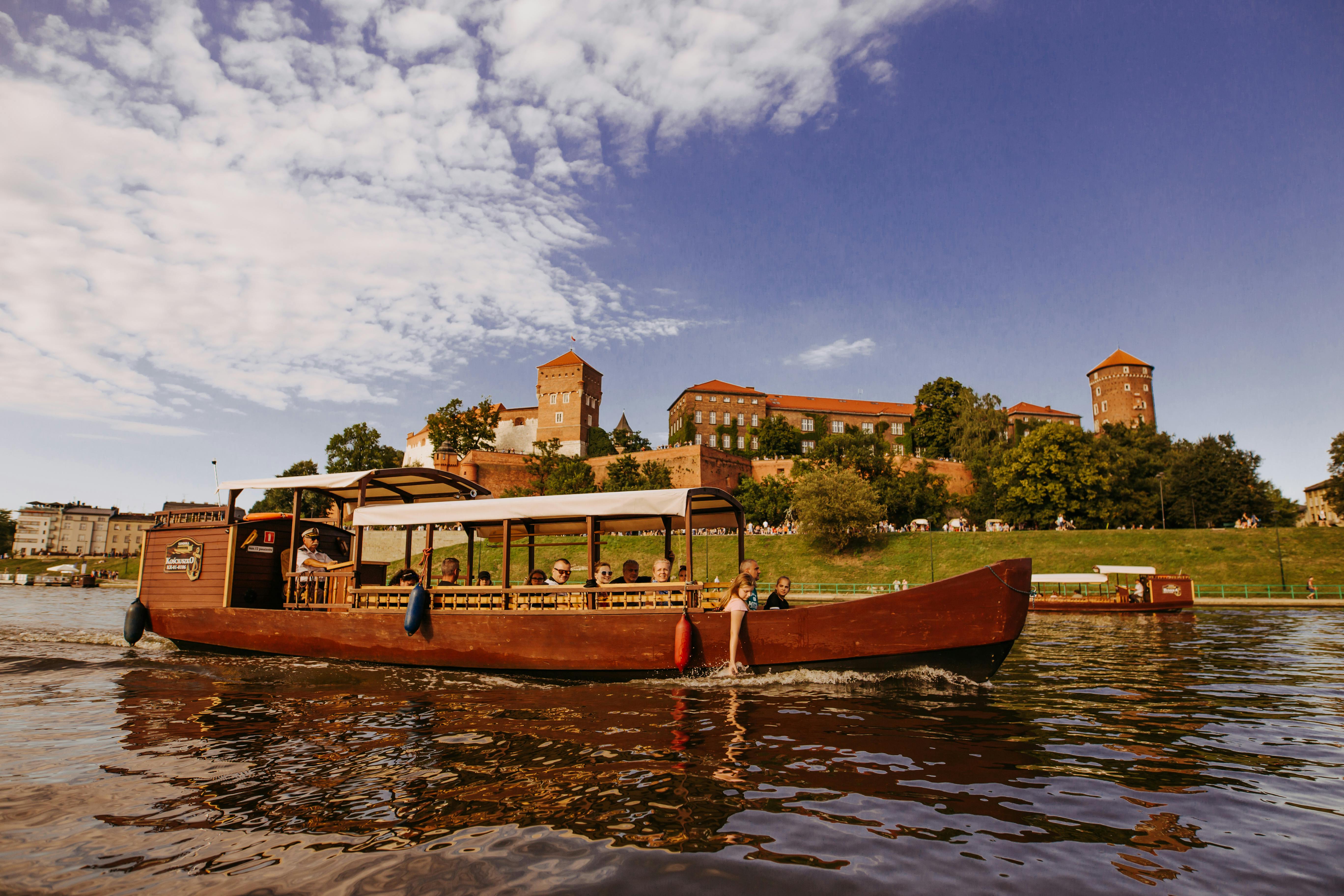 A wooden tour boat with passengers is on a river; in the background, a historic building and green hillside under a partly cloudy sky.