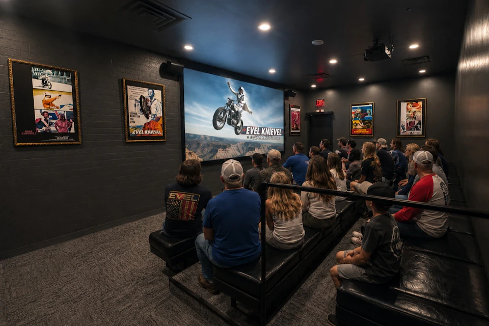 A group of people seated in a small theater, watching a projected image of Evel Knievel performing a motorcycle jump.