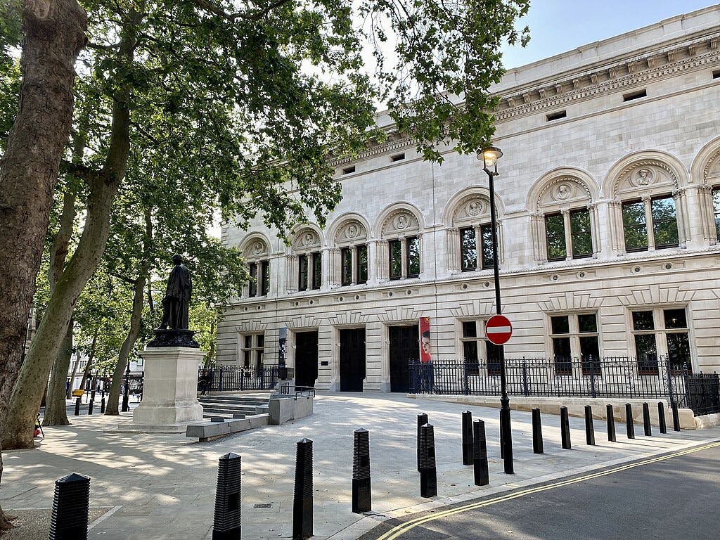 Grand bâtiment en pierre avec des fenêtres cintrées, une statue, des arbres et des bornes noires. Un panneau d'interdiction d'accès est visible.