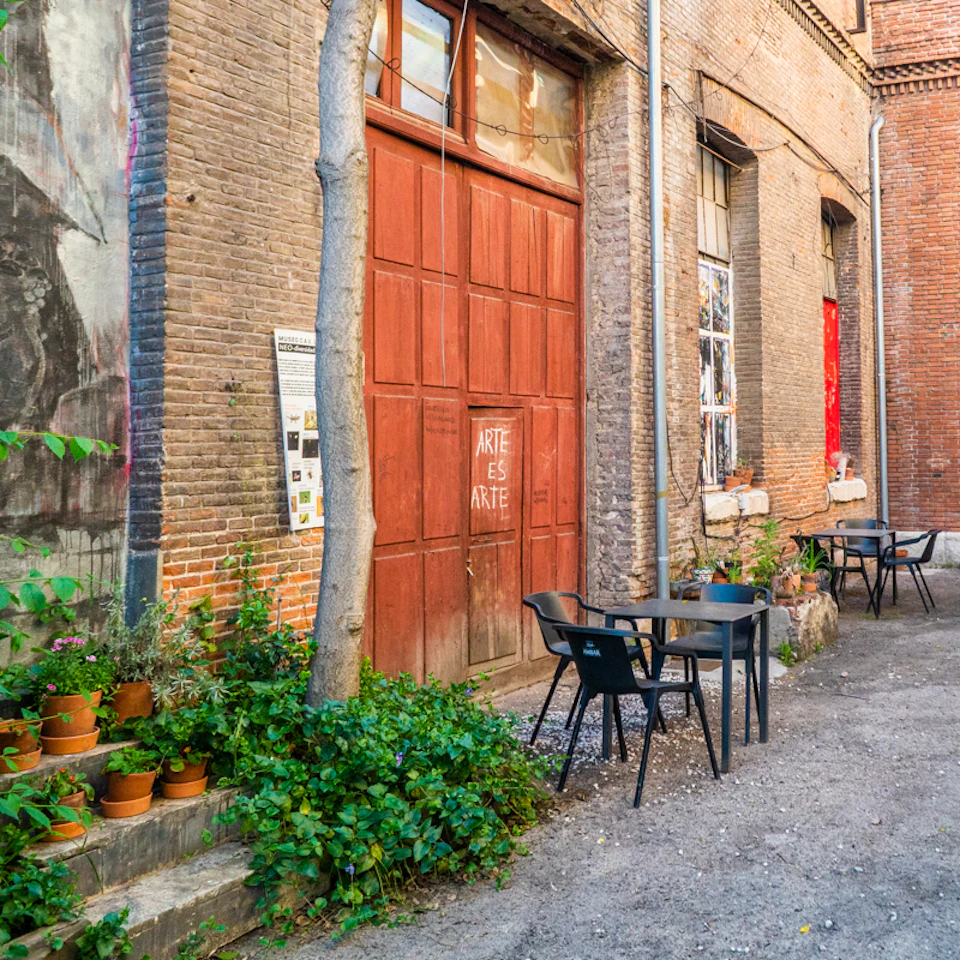 An alley with a red wooden door, potted plants, a tree, and outdoor tables and chairs against brick walls. "Arte Es Arte" is written on the door.