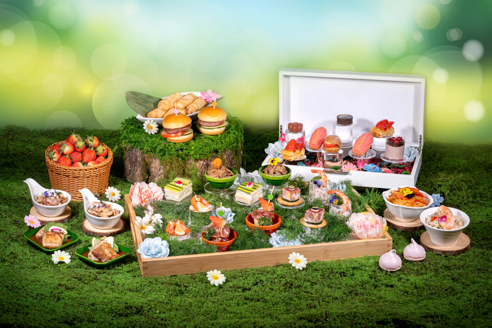 An elaborate display of food items including pastries, burgers, and desserts arranged on a grassy surface with floral decor.