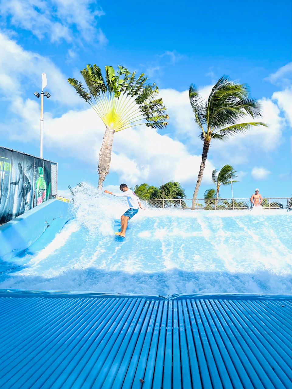A person surfs on a blue artificial wave under a bright, clear sky with palm trees swaying in the background.