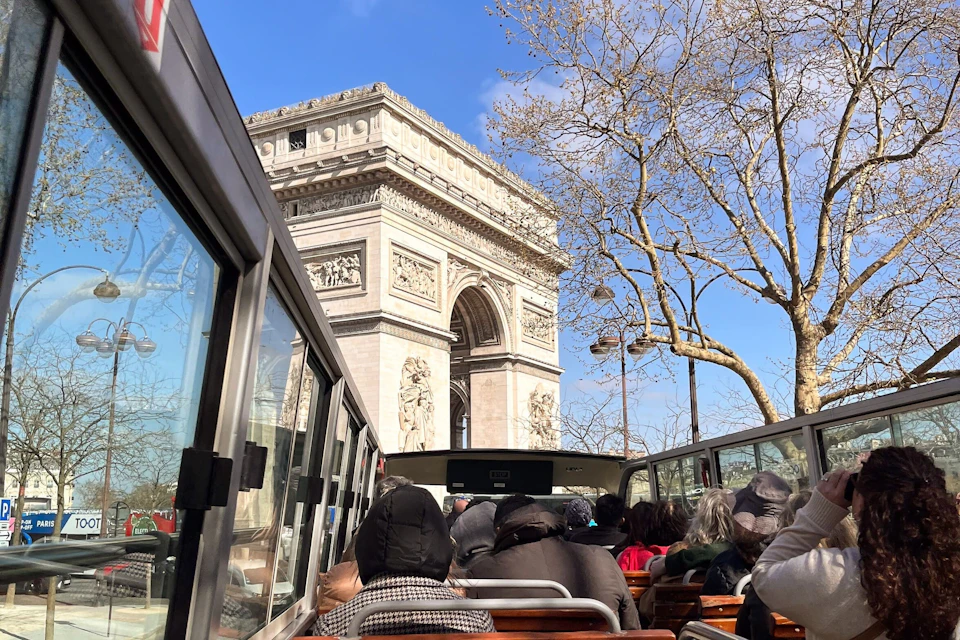 Tourists sitting on an open-top bus near the Arc de Triomphe, with trees and a clear blue sky in the background.