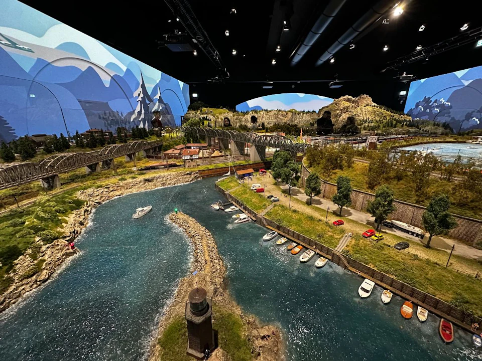 Detailed model landscape with a river, boats, cars, and bridges, set against a mountainous backdrop under a ceiling with lights.