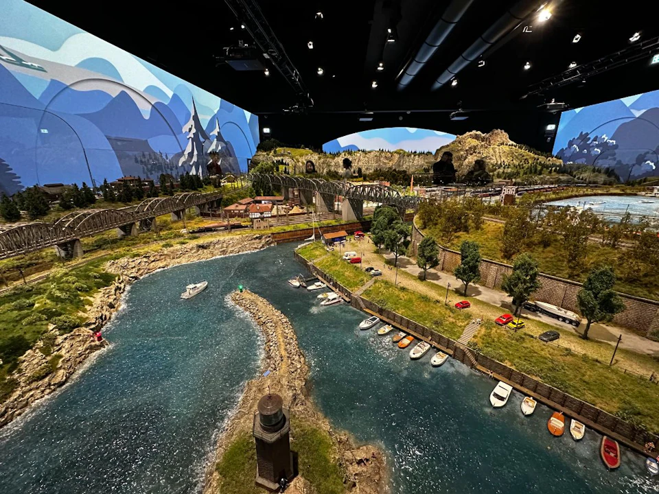 Detailed model landscape with a river, boats, cars, and bridges, set against a mountainous backdrop under a ceiling with lights.