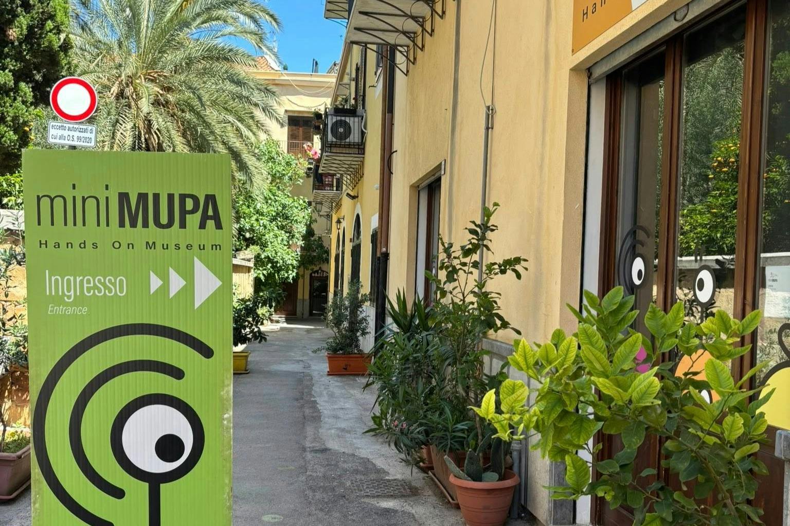 狭窄的小巷，盆栽植物，绿色标志“MUPA”指向右侧，晴朗的天空下有黄色建筑。