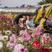 Un couple dans un véhicule de camouflage au milieu d'un champ de fleurs coloré, prenant un selfie et souriant.