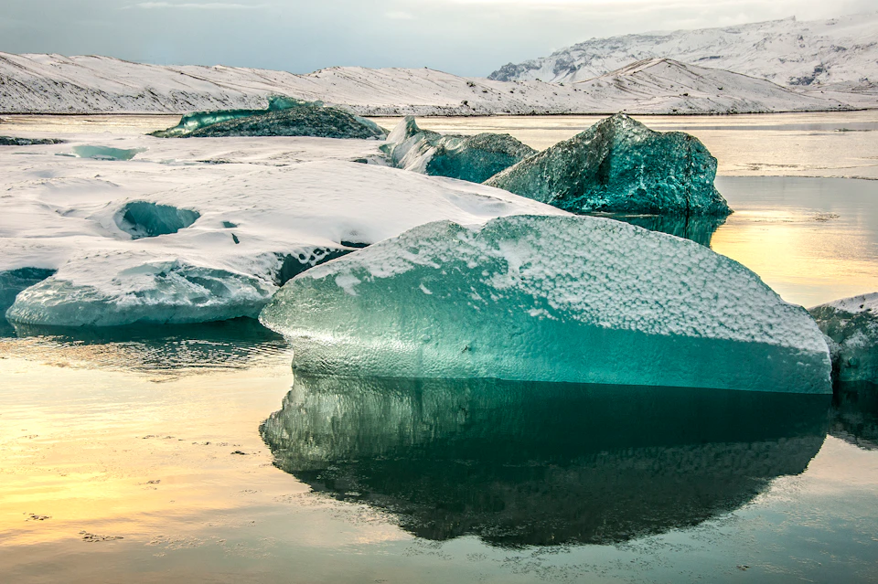 Large chunks of blue and white ice float on a calm body of water, with snow-covered mountains in the background.