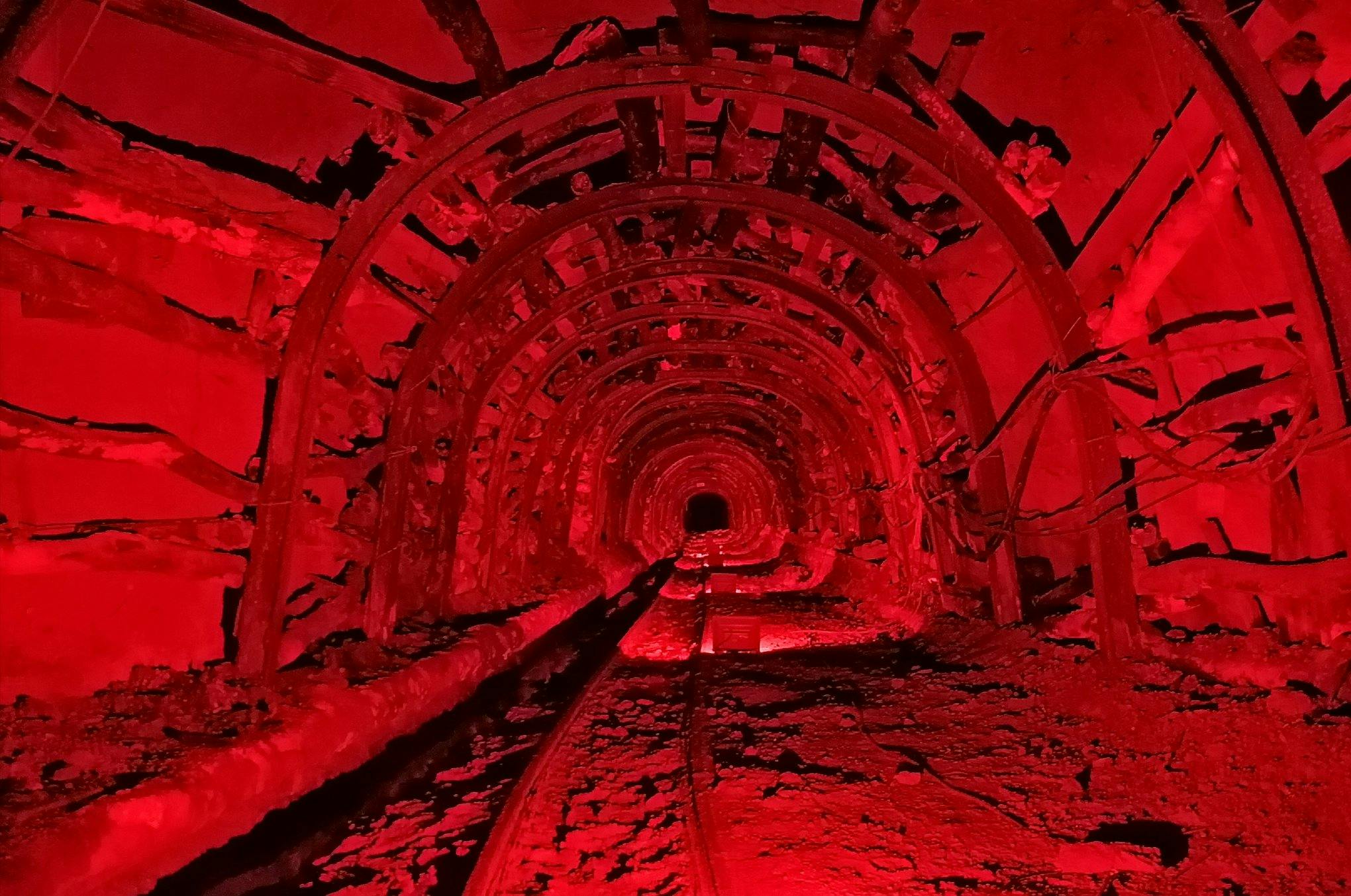 A dimly lit underground tunnel with red lighting, arched metallic supports, and tracks running along the floor.