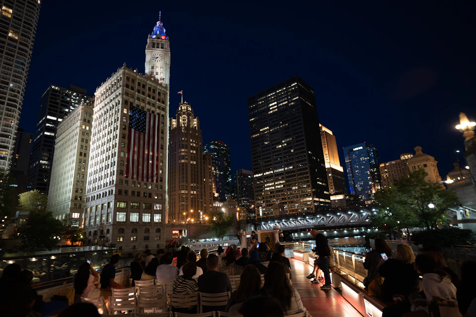 People seated on a boat at night, with illuminated skyscrapers and a bridge over a river in the background.