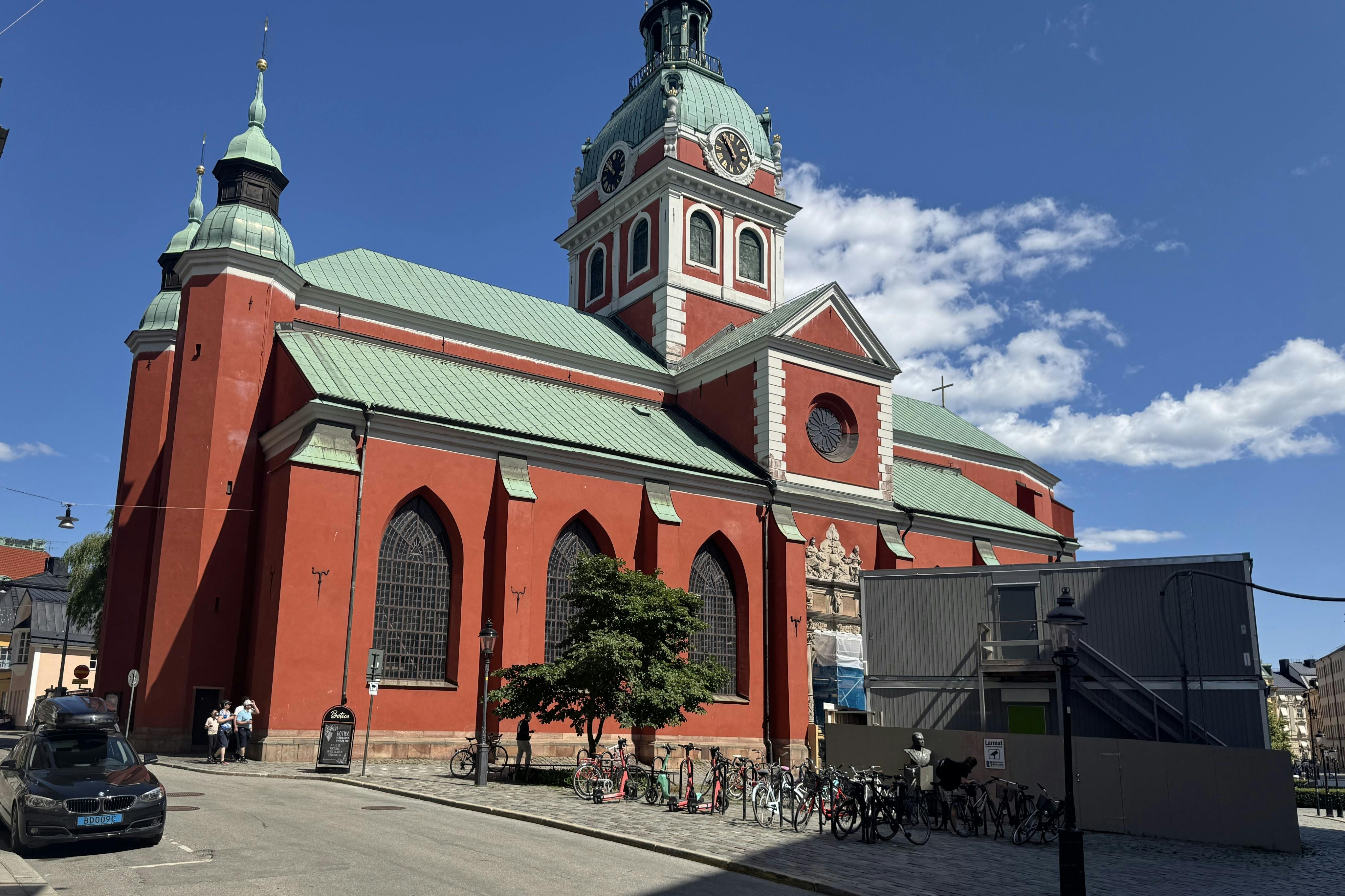 A red and green-roofed church with a clock tower under a blue sky, surrounded by a few bicycles and pedestrians.