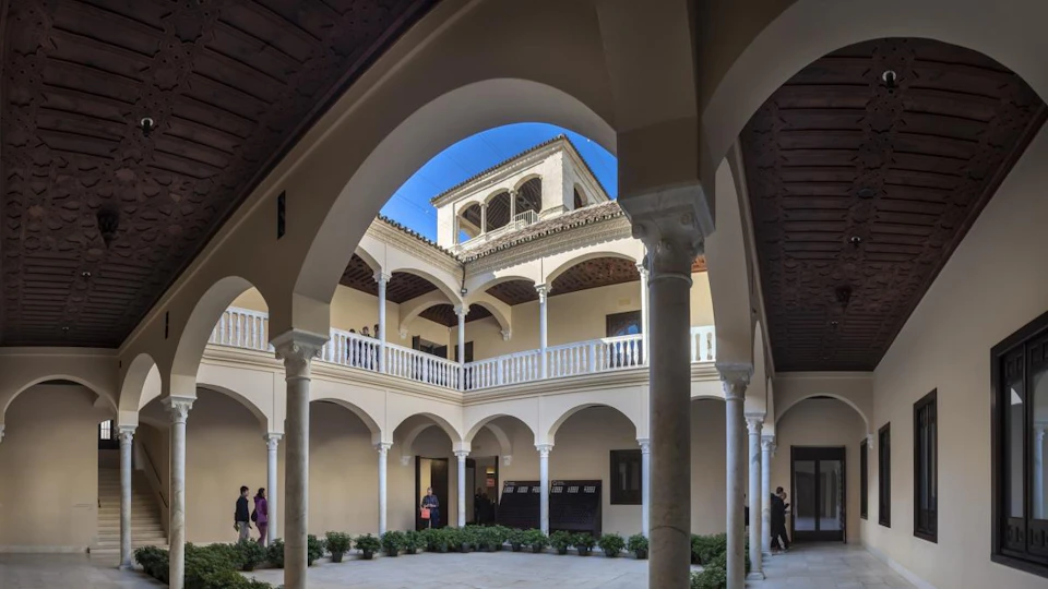 A courtyard with arched columns, an upper balcony, potted plants around the perimeter, and people walking and standing below.