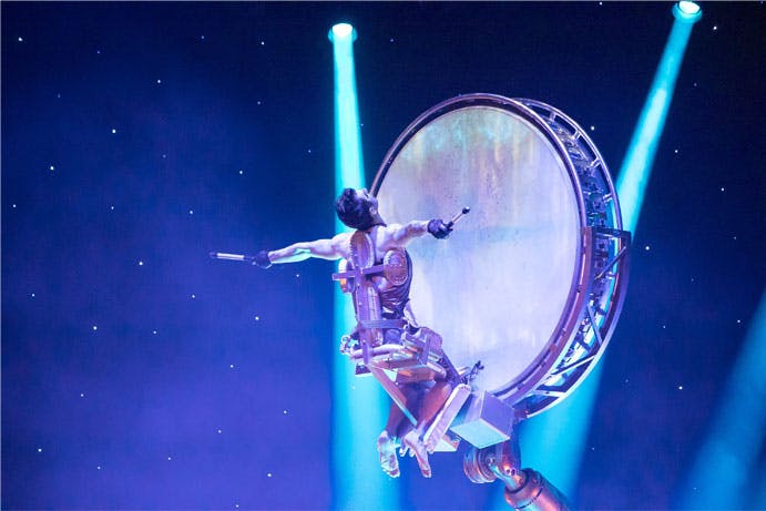 Účinkující ve futuristickém obleku hrající na velkém rotujícím bubnu na jevišti s modrými reflektory a hvězdným pozadím.