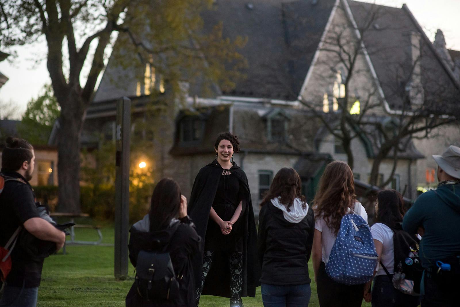 A person in a black cape stands and smiles outdoors, surrounded by four others, with a large building in the background.