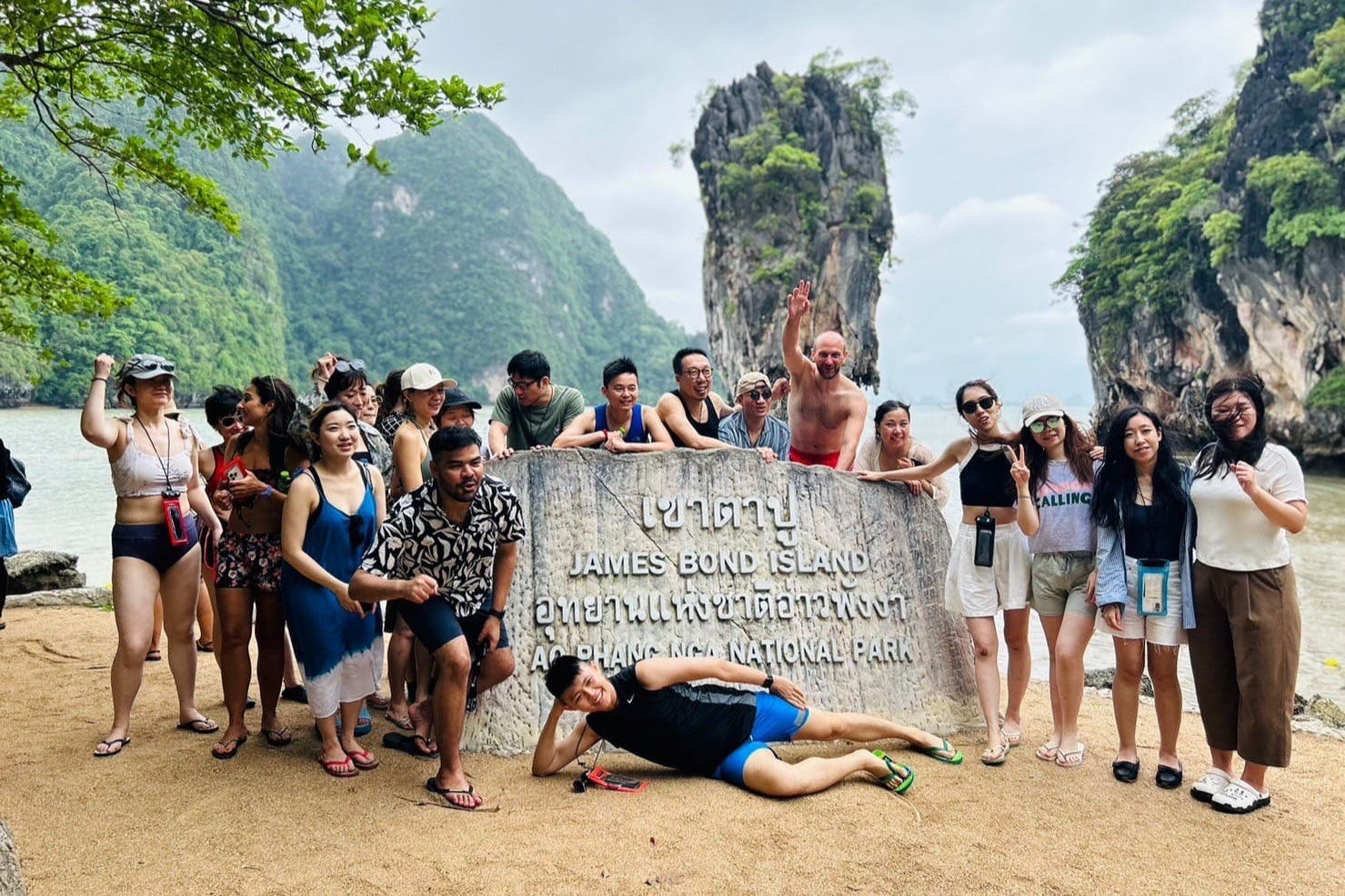 Skupina lidí pózuje kolem vícejazyčného nápisu na ostrově Jamese Bonda s velkými skalními útvary a zelení v pozadí.