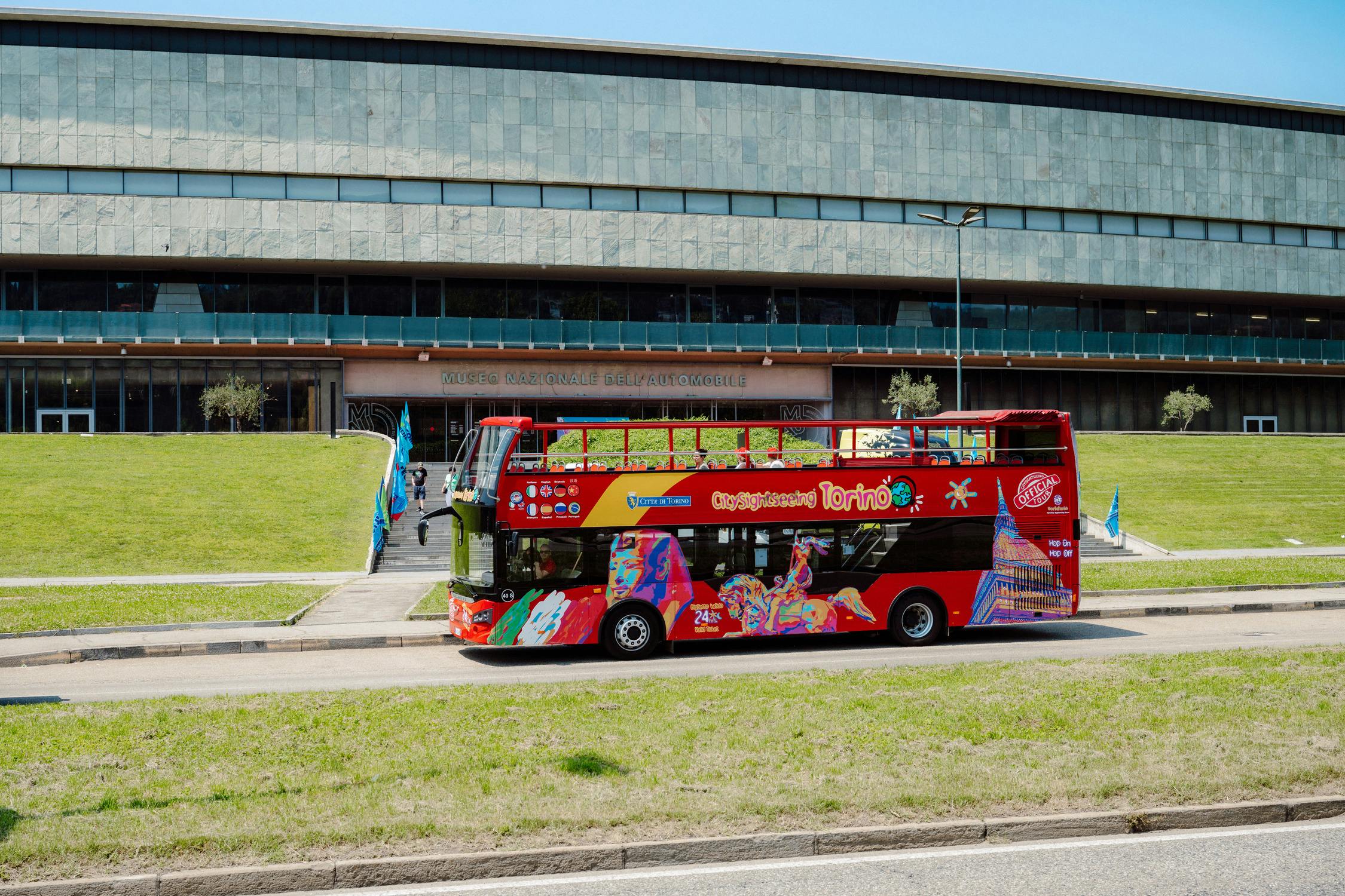 A red double-decker tour bus parked in front of a modern building with a grassy lawn and steps leading to the entrance.