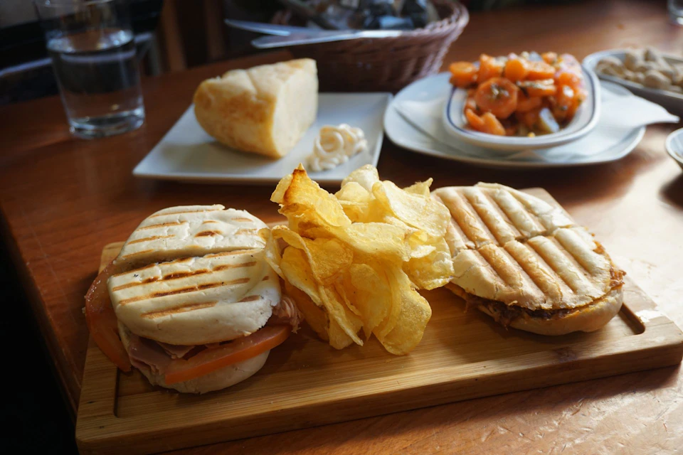 A meal consisting of two panini sandwiches, potato chips, a plate with salad, and a block of cheese on a wooden table.