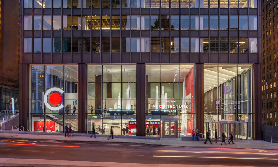 The exterior of the Chicago Architecture Center, showcasing large glass windows with architectural models inside, and people walking by.