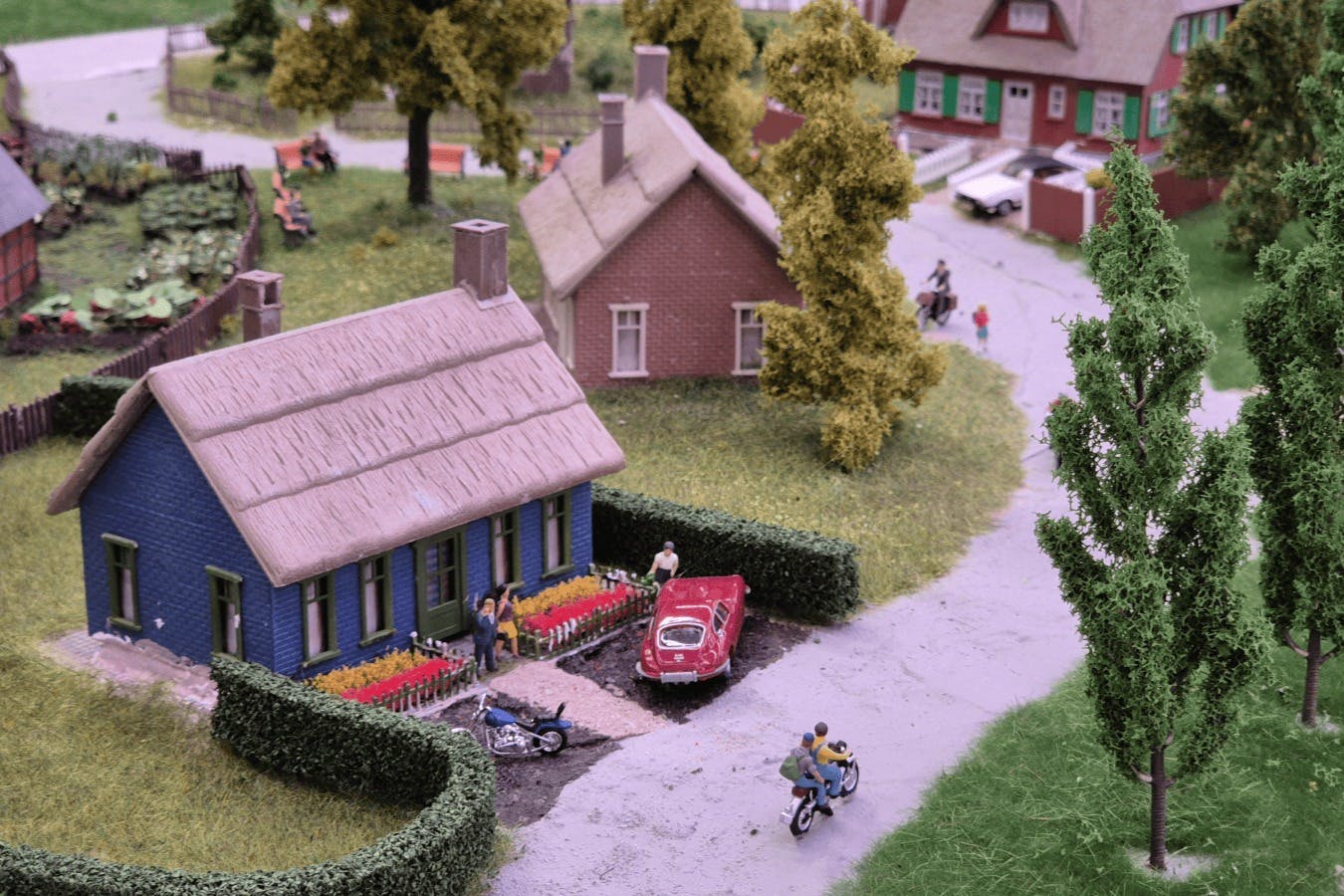 A detailed model of a village scene with houses, cars, motorcycles, trees, and miniature people on a winding road.