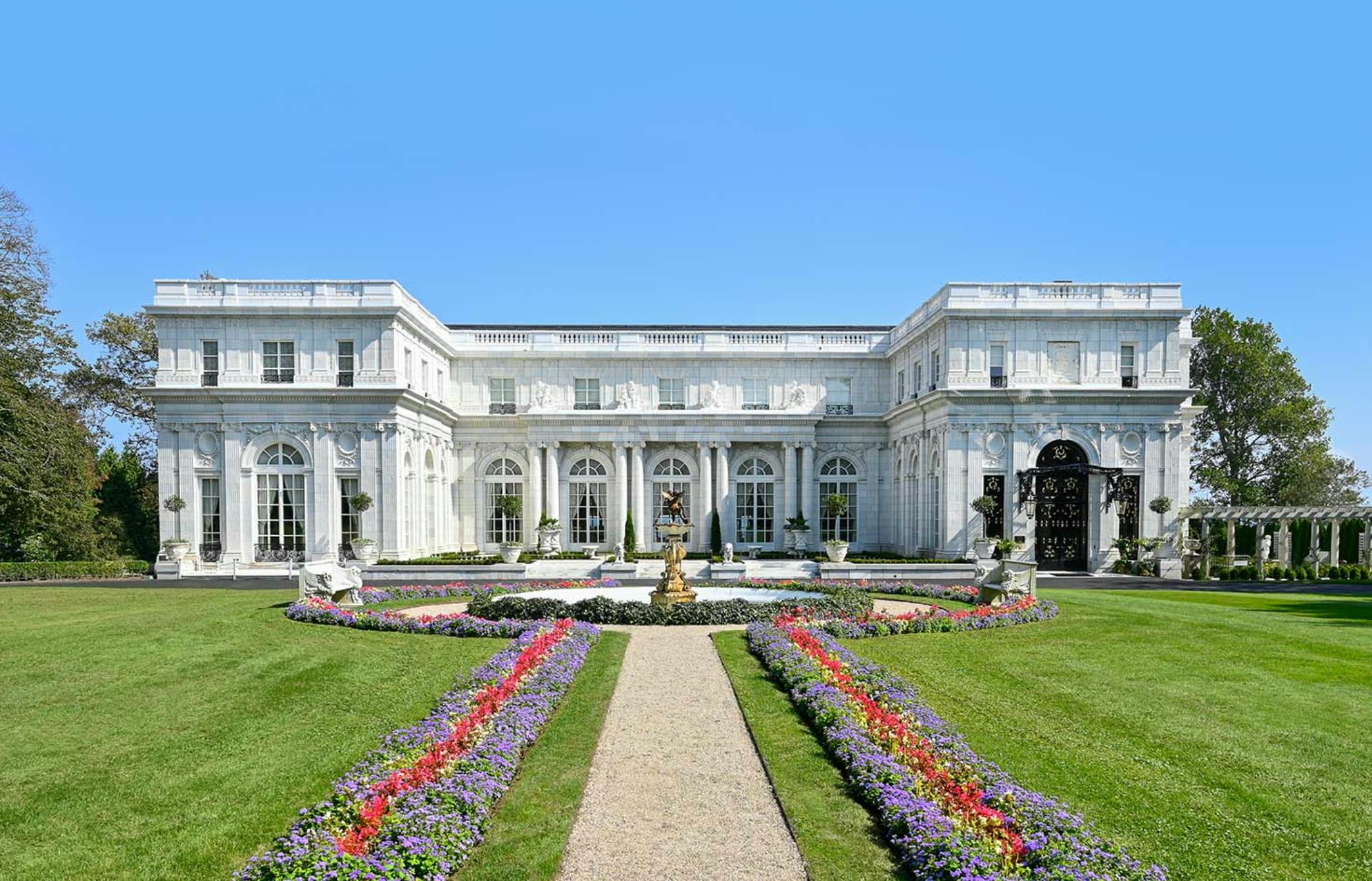 Um imponente edifício branco com janelas em arco e colunas, uma fonte e um jardim de flores coloridas em primeiro plano.